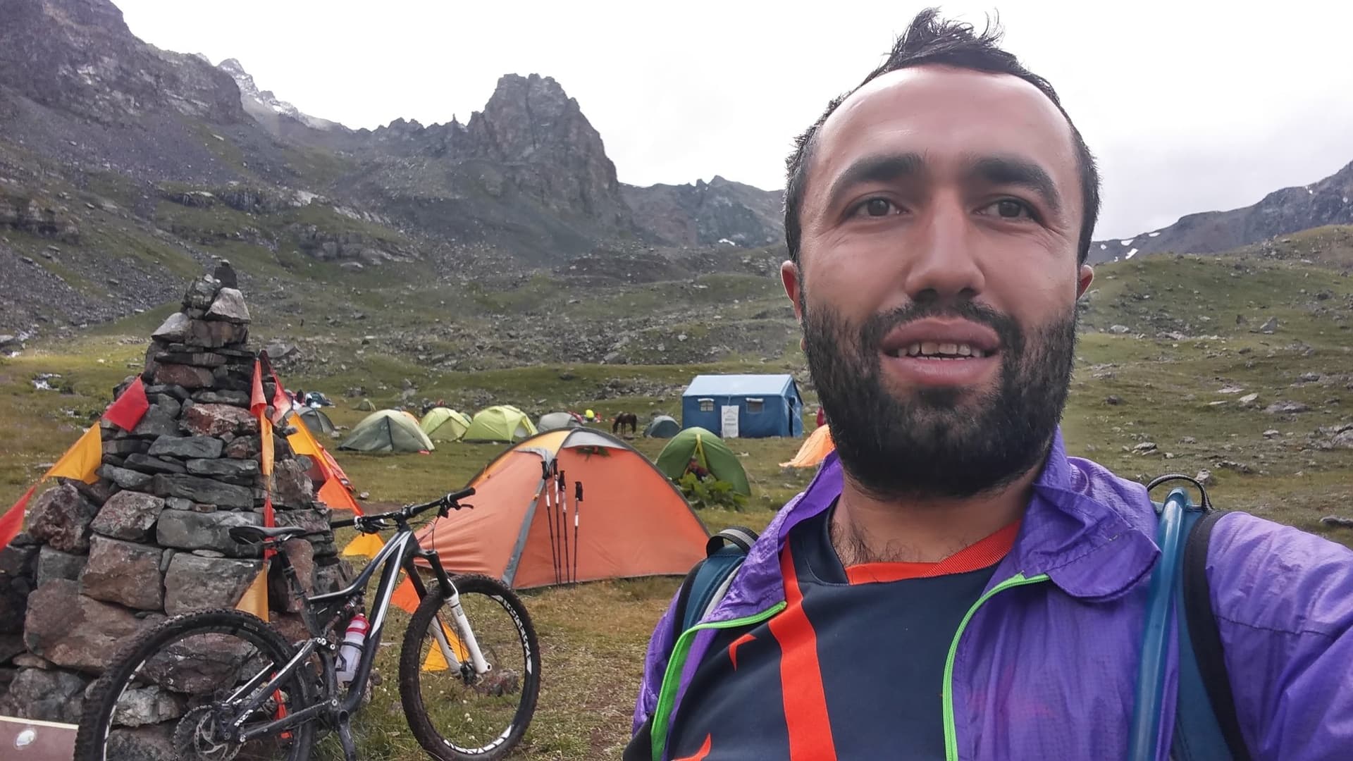 Camper posing with a mountain bike and tents pitched beneath jagged peaks in the Kackar Mountains