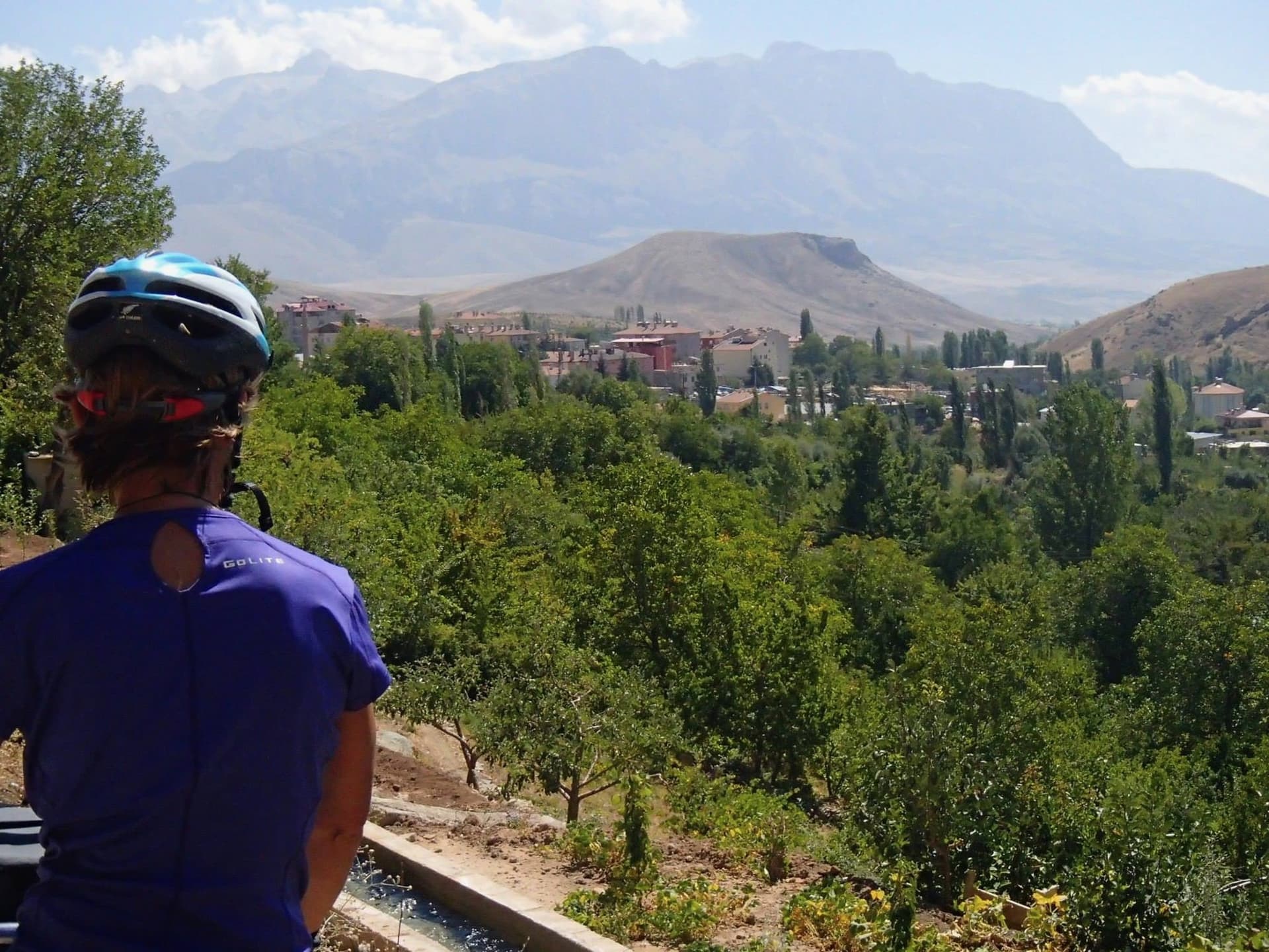 Cyclist pausing above a green valley of orchards and rooftops in Cappadocia
