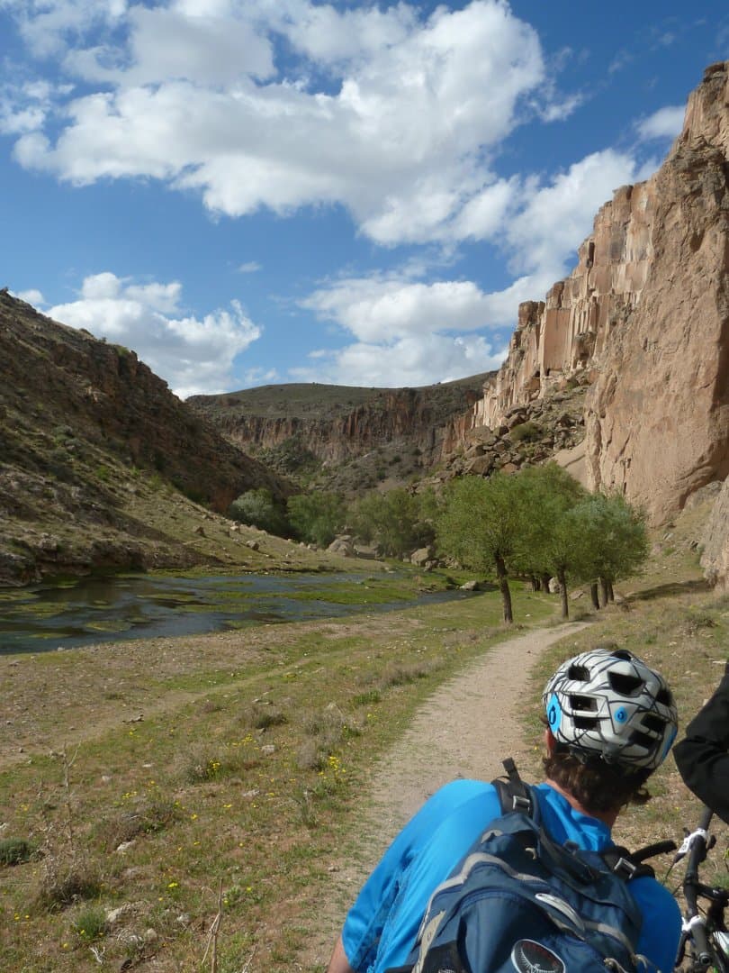 Cyclist riding a narrow trail beside a river lined with cliffs and trees