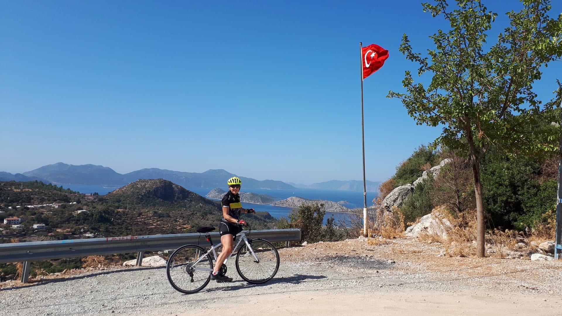 Cyclist standing beside a Turkish flag on a hillside above the Aegean coast