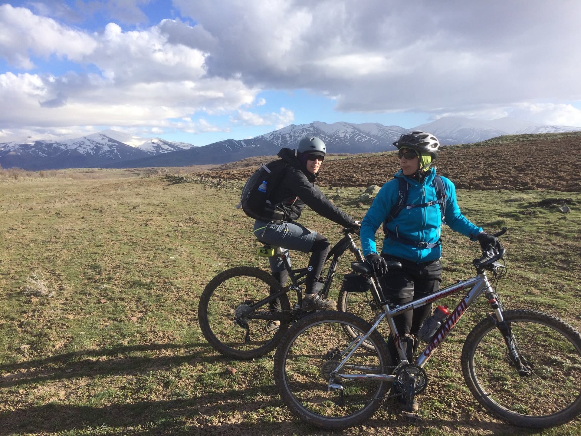 Cyclists with their bikes on a grassy hillside below snow-capped mountains