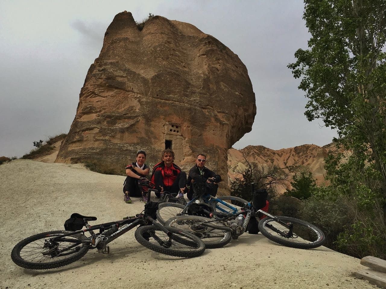 Cyclists resting with their bikes beneath a massive balanced boulder in Cappadocia