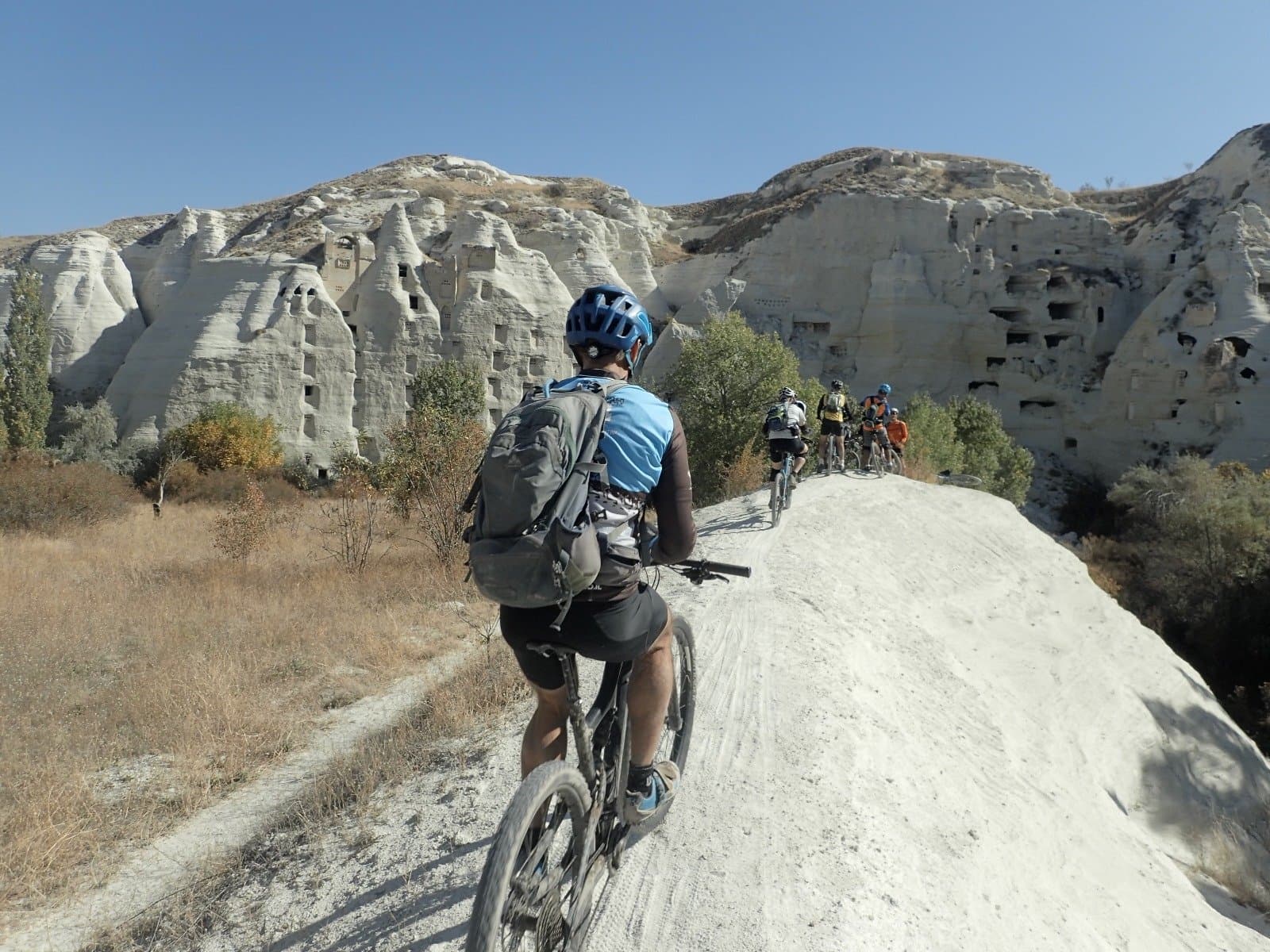 Cyclists riding a pale rock trail past cave dwellings carved into a Cappadocia cliff