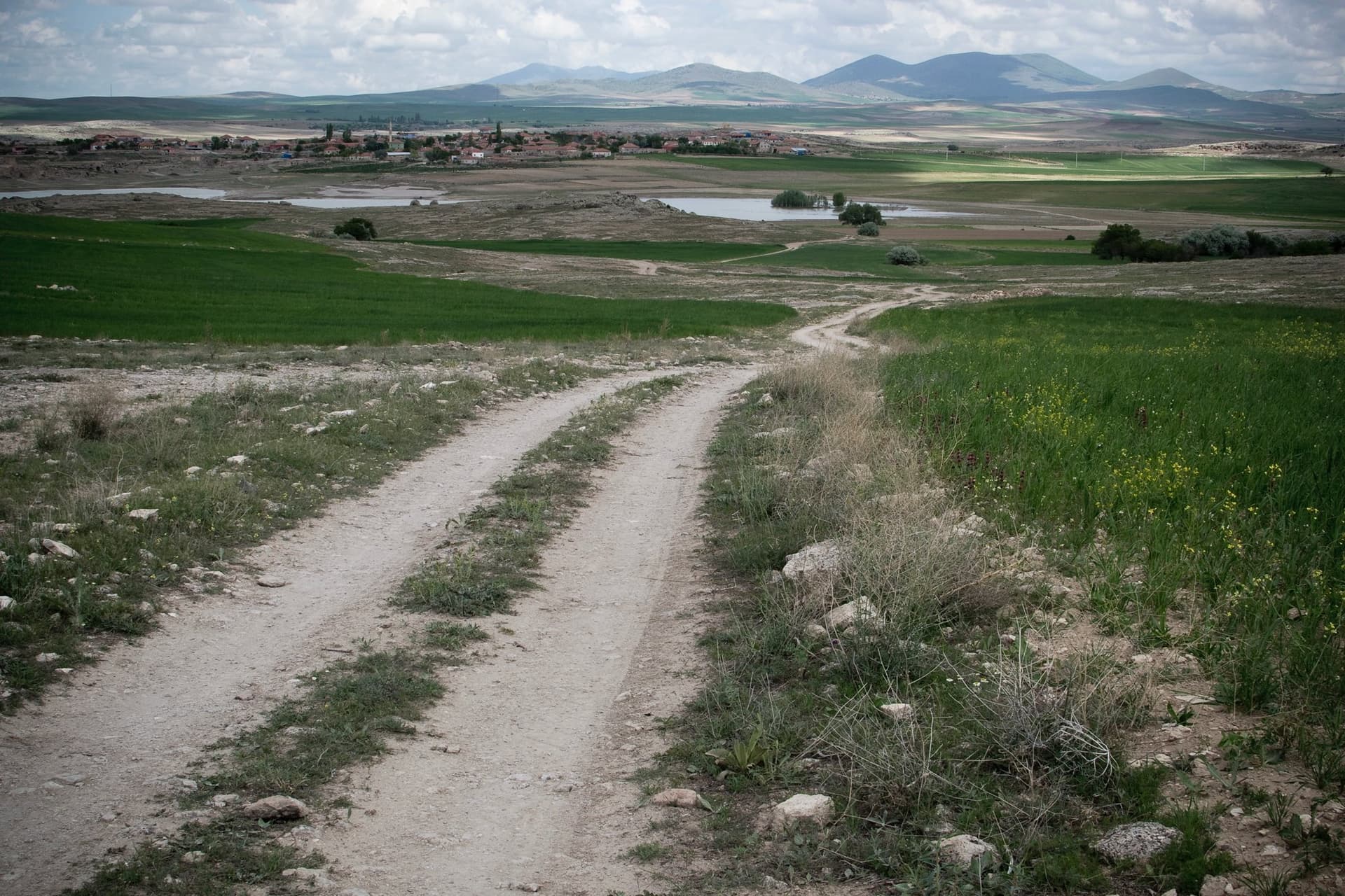 Dirt track running past green fields toward a distant lake and village