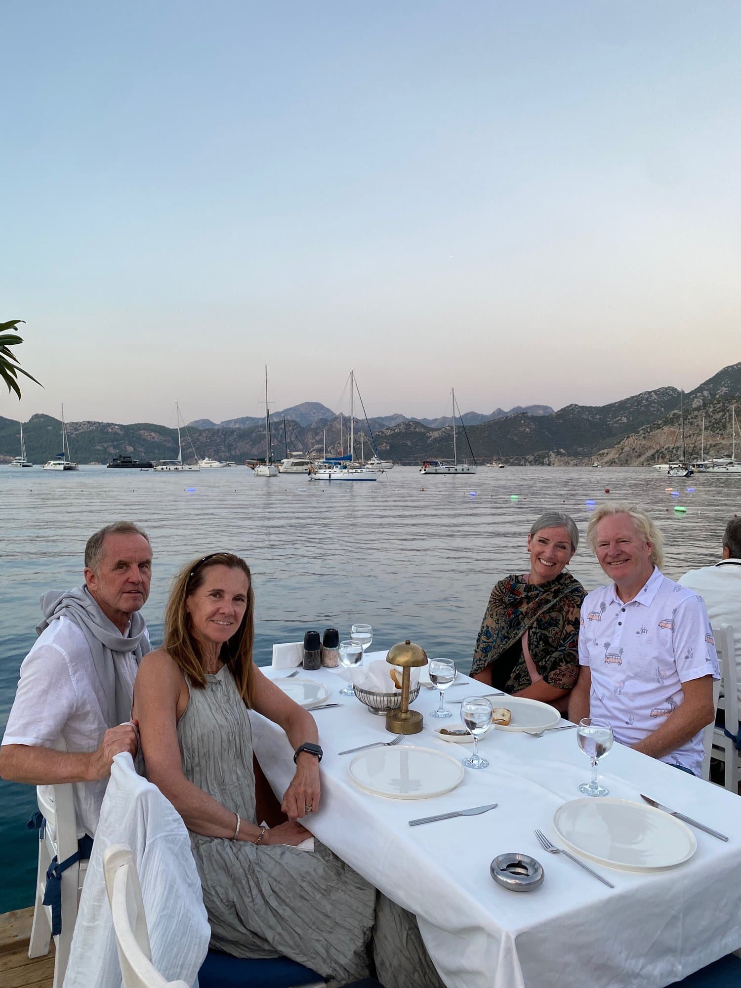 Group dining at a waterside table overlooking moored yachts at dusk on the Aegean coast