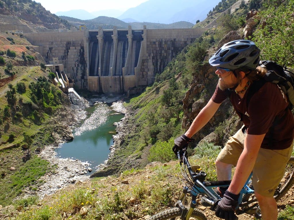 Mountain biker beside a rocky stream below a large dam wall in the Atlas Mountains