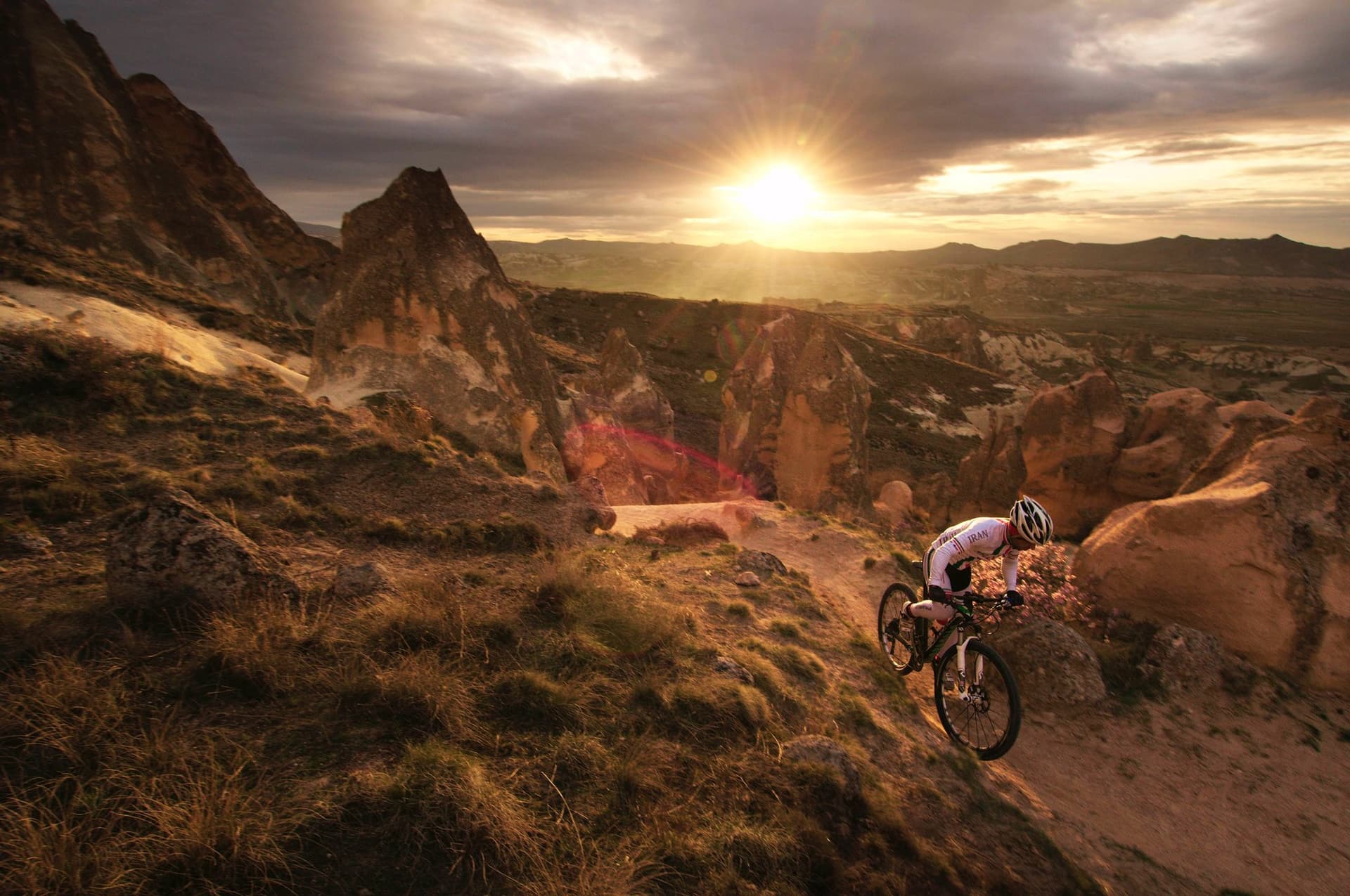 Mountain biker descending a red valley trail in Cappadocia under a dramatic sunset sky