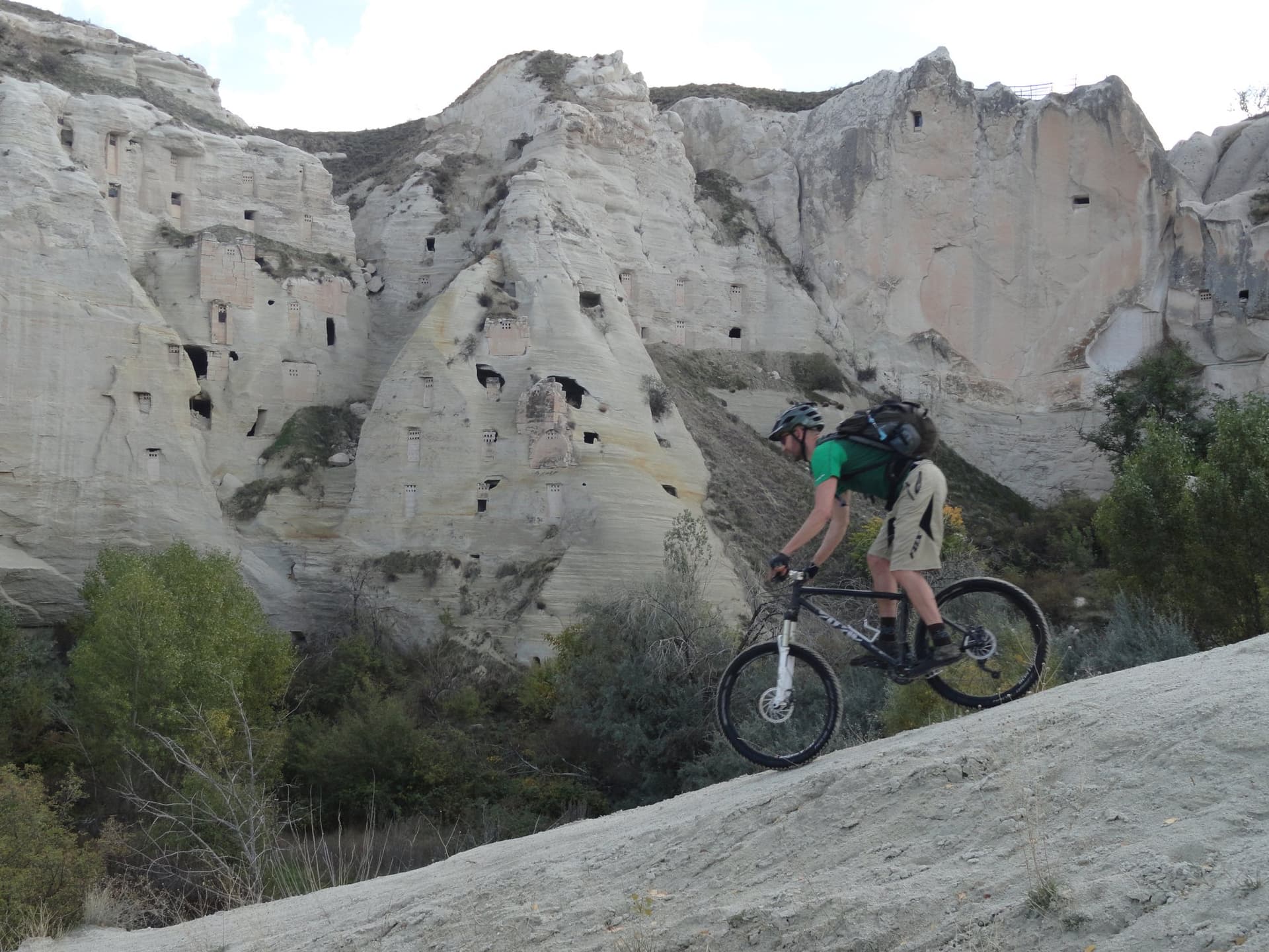 Mountain biker riding a steep pale slope below cave-riddled tuff cliffs in Cappadocia