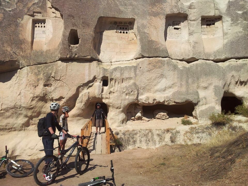Mountain bikers with their bikes below cave dwellings carved into a Cappadocia cliff