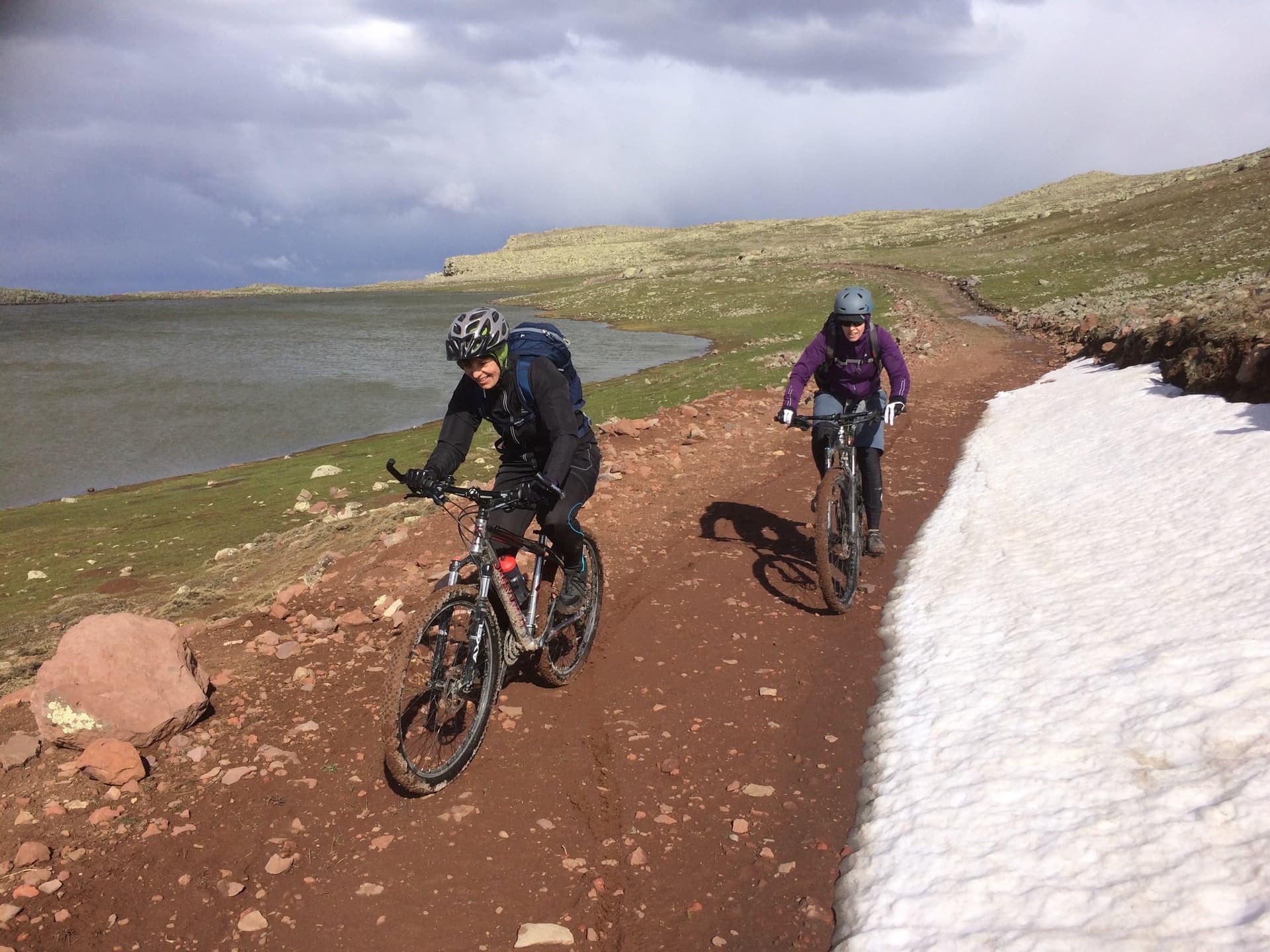 Mountain bikers riding a red-earth trail beside snow patches near a lake