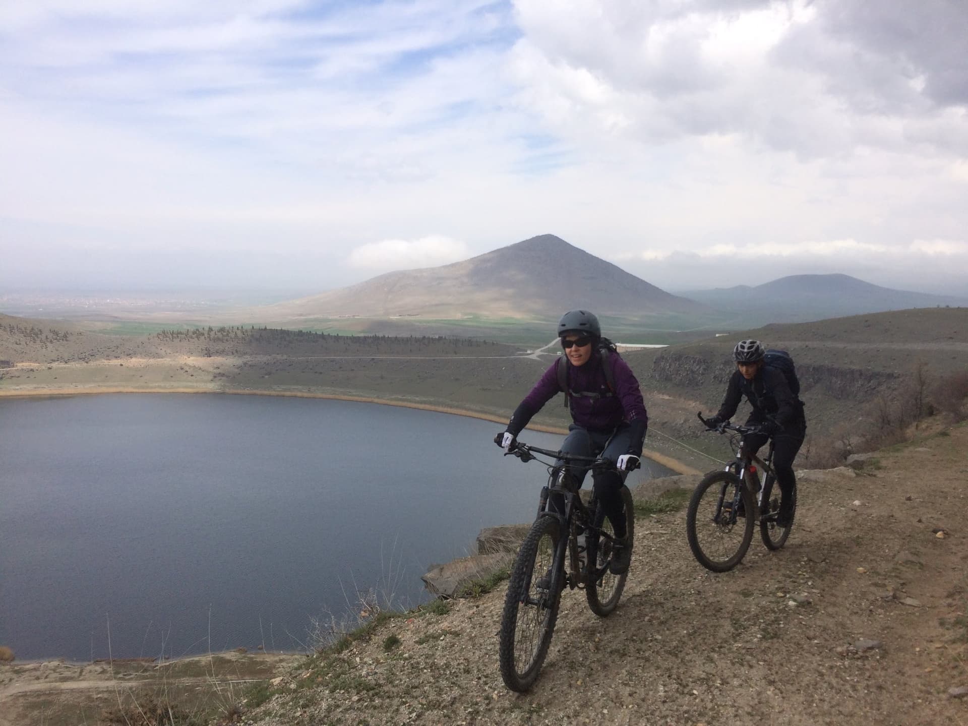 Mountain bikers riding a ridge trail above a still lake with a hill beyond