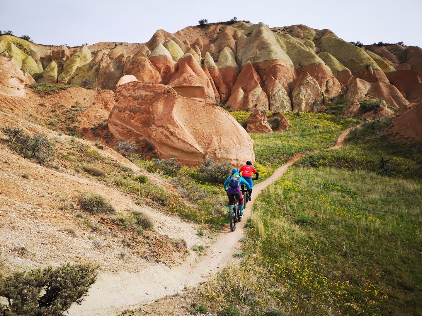 Mountain bikers riding a trail beneath layered red and yellow rock formations in Cappadocia