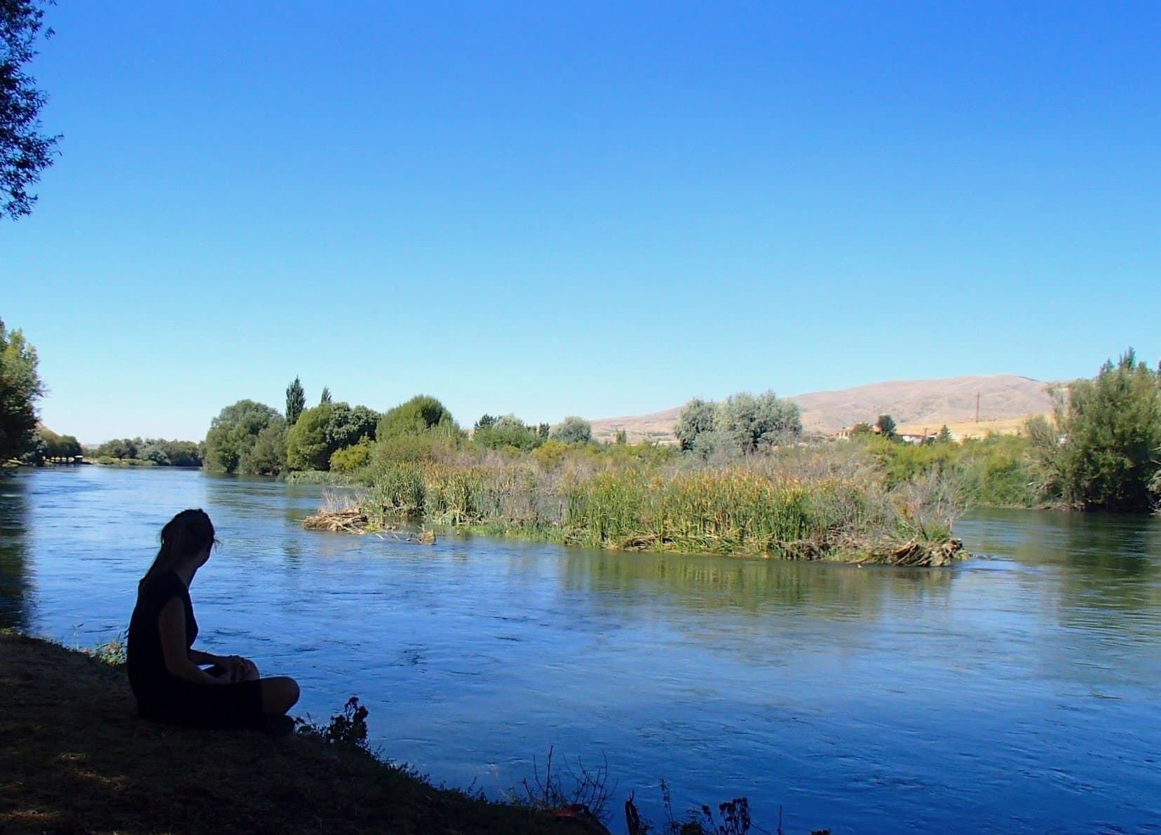 Person sitting quietly on a riverbank beside a wide green river