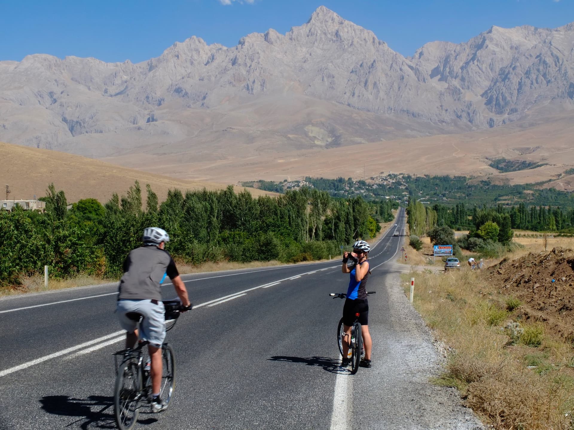 Two road cyclists on a straight valley road heading towards snow-capped mountains