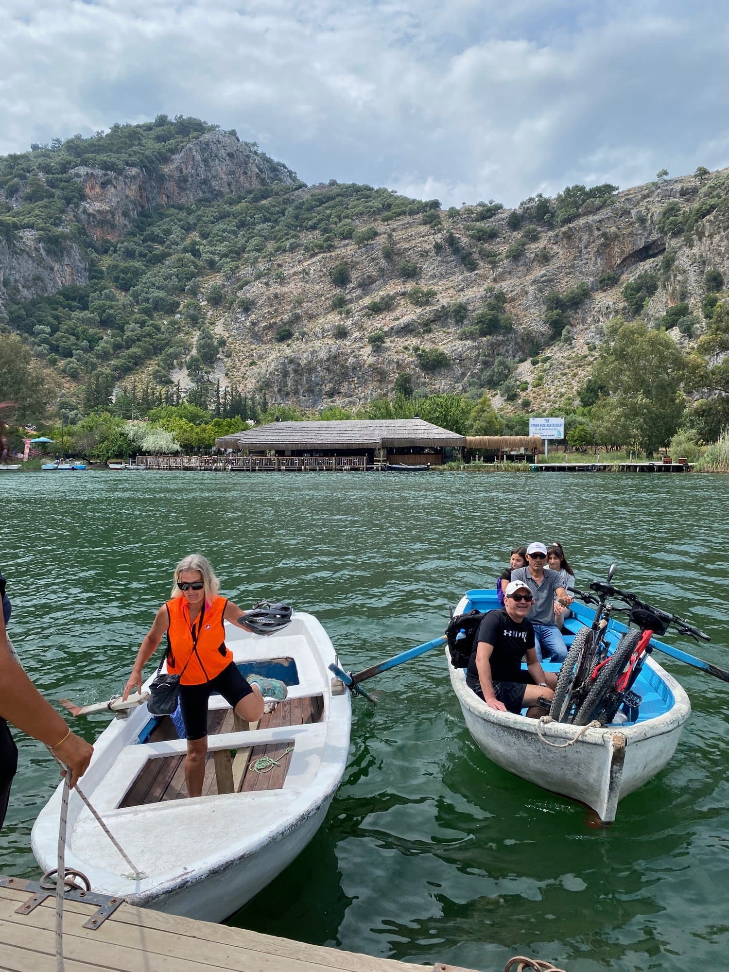 Two rowboats loaded with bicycles crossing a lagoon toward a lakeside restaurant on the Aegean coast