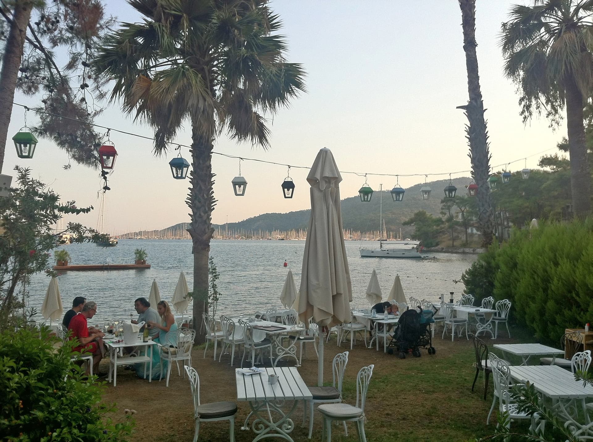 Waterfront restaurant tables under palm trees strung with lights at dusk